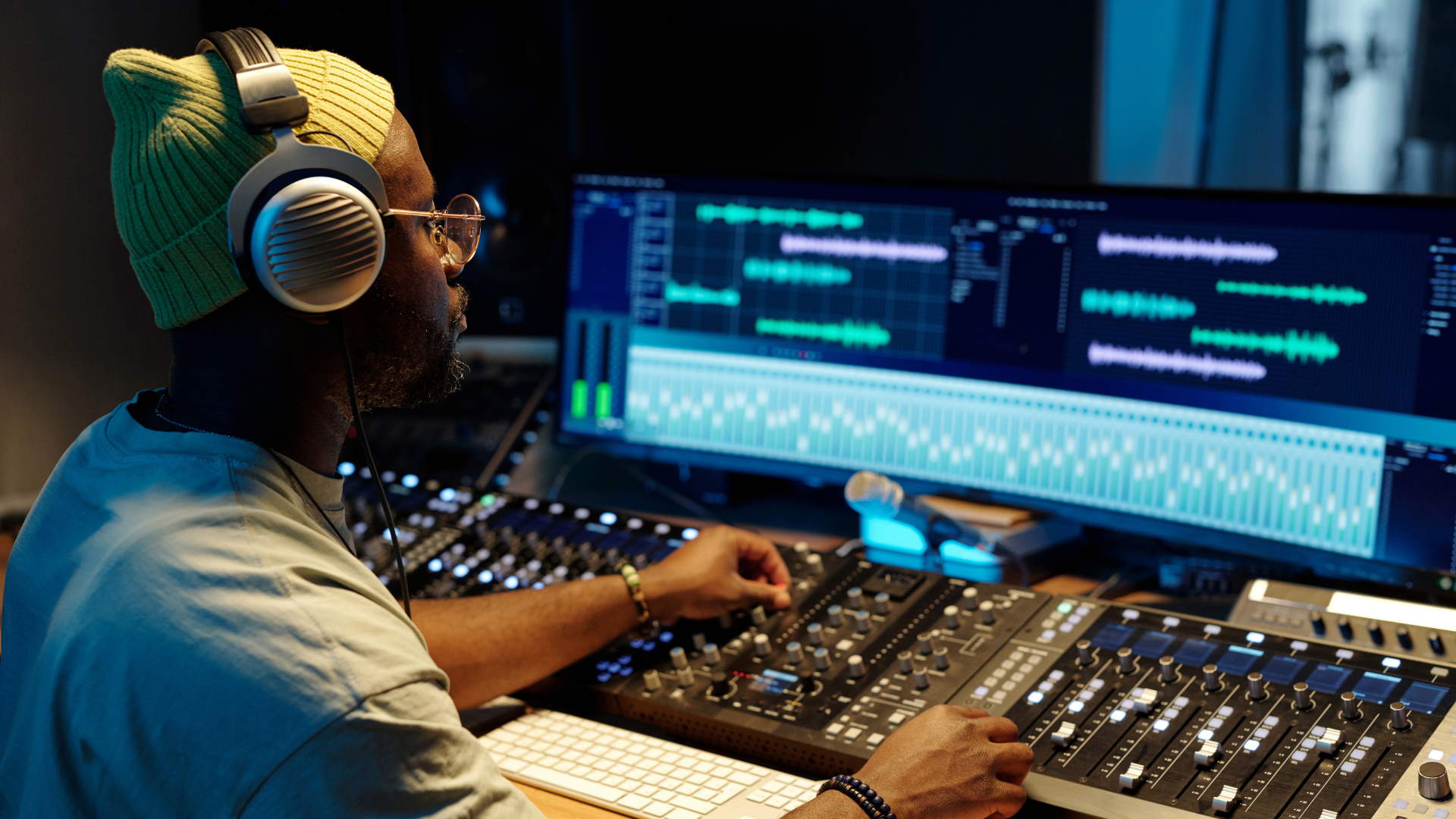 A person wearing headphones works at a sound mixing console in front of a wide monitor displaying audio waveforms.