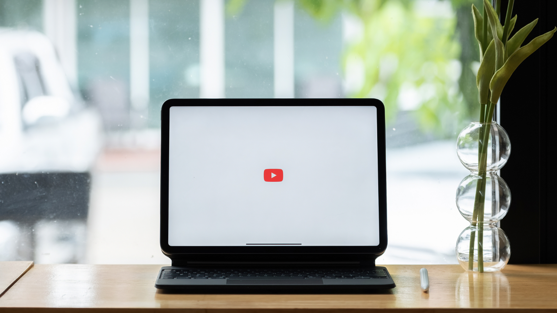 A tablet with the YouTube logo on its screen, resting on a wooden desk next to a vase of greenery in front of a window.