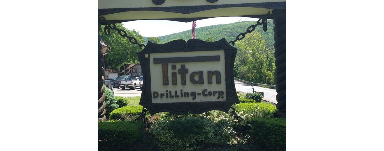 Sign for Titan Drilling-Corp, hanging by chains, set in front of foliage with a mountain in the background.