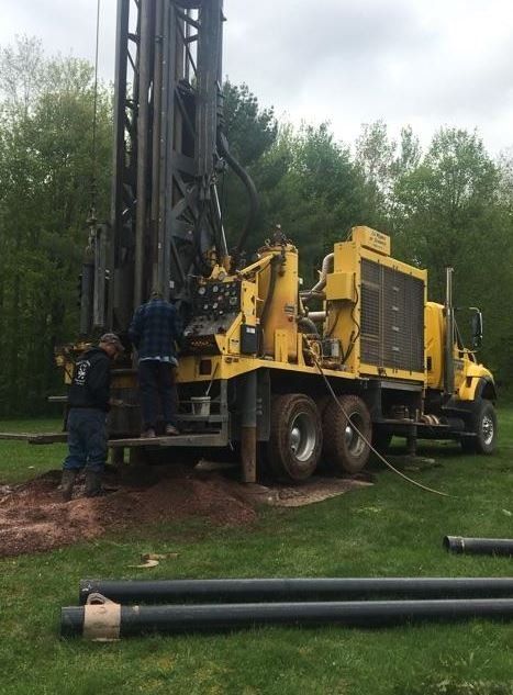 Yellow drilling rig on green grass; two workers beside it. Black pipes lie in the foreground.