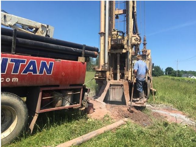 A drilling rig beside a red truck, worker at the rig on a grassy field on a sunny day.