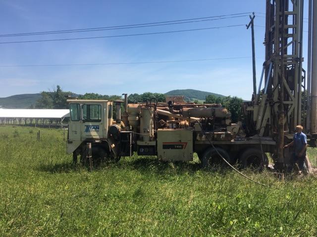 A drilling rig in a grassy field with a worker, rural landscape and clear blue sky in background.