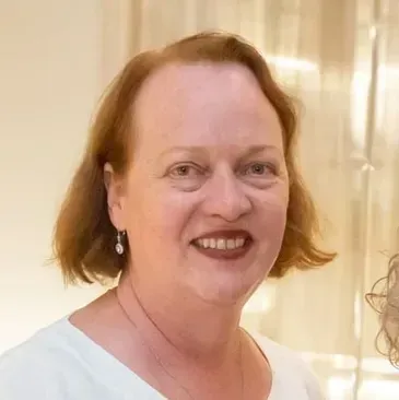 A smiling person with shoulder-length reddish-brown hair, wearing a white top and small drop earrings.