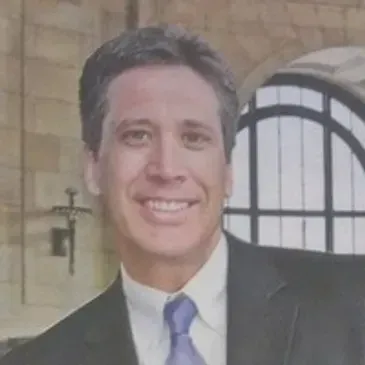 A smiling person in a dark suit and blue tie posing in front of a stone wall and arched window.