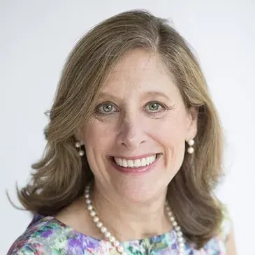 A smiling woman with shoulder-length wavy brown hair wearing a floral top and a pearl necklace against a white background.