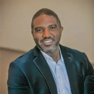 A smiling person wearing a teal velvet blazer and a light-colored button-down shirt against a neutral background.