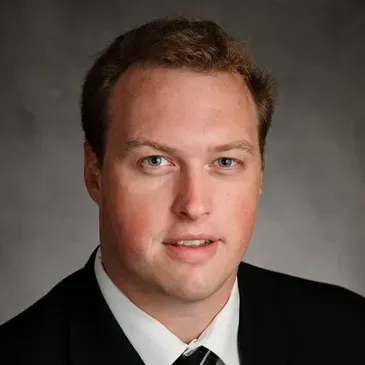 A headshot of a person wearing a black suit and white collared shirt against a gray background.