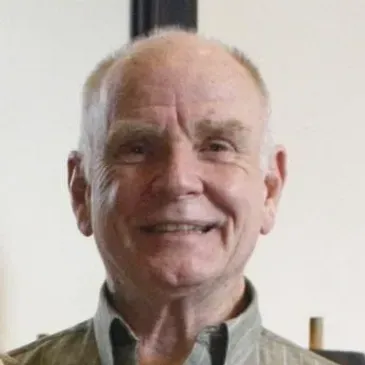 A person with light skin and short hair smiling warmly against a plain, light-colored background.