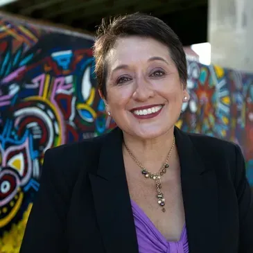 A smiling person in a black blazer and purple top posing in front of a colorful, abstract patterned mural.