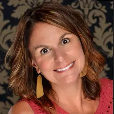 A smiling person with shoulder-length brown hair, wearing long gold earrings and a textured red top against damask wallpaper.