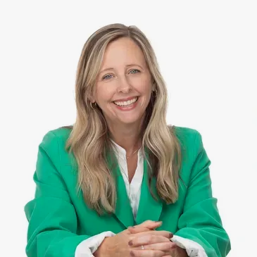 A smiling woman with long blonde hair, wearing a green blazer over a white shirt, resting her hands on a surface.