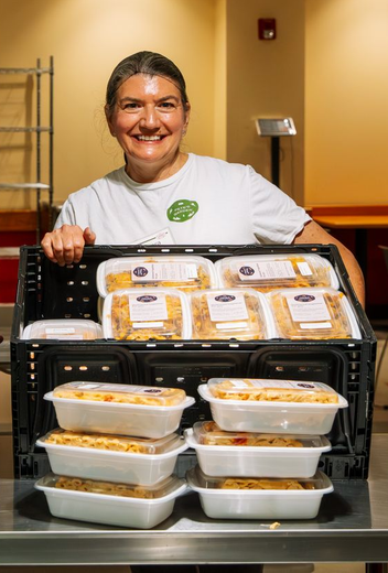 A smiling person in a white t-shirt stands behind a black crate filled with several packaged food containers.
