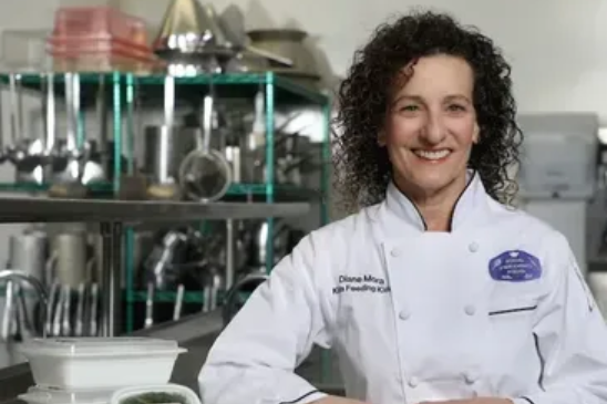 A chef wearing a white uniform smiles in a professional kitchen with stainless steel equipment in the background.