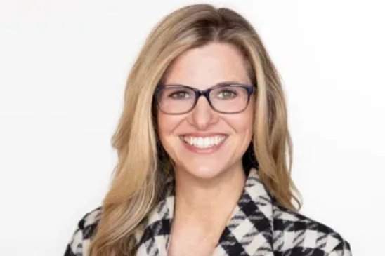 A smiling person with long blonde hair and glasses, wearing a black and white patterned blazer against a white background.