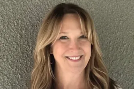 A person with long, layered blonde hair and hoop earrings smiling warmly against a textured, light gray background.