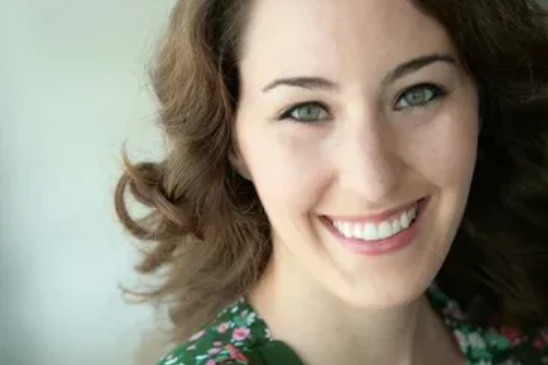 A close-up portrait of a smiling person with wavy brown hair, wearing a green floral-patterned top.