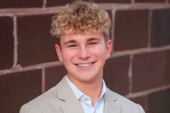 A smiling person with curly light hair, wearing a beige blazer and white collared shirt, against a brown brick wall.