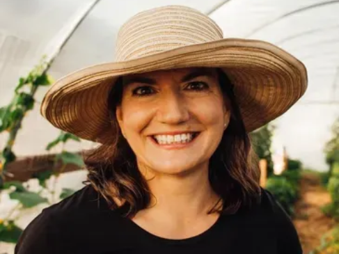 A smiling person wearing a wide-brimmed straw hat inside a greenhouse with plants in the background.