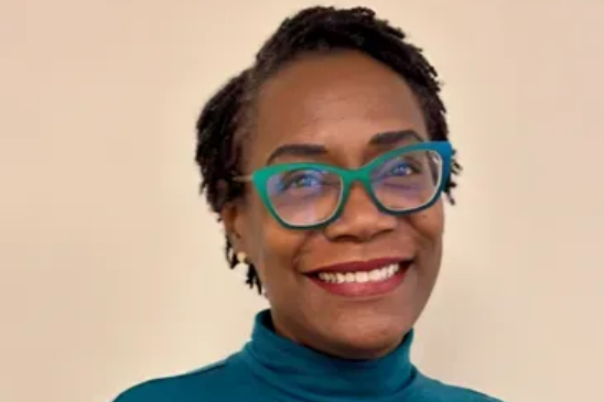 A person with short, dark, textured hair wears teal glasses and a teal turtleneck, smiling against a neutral background.