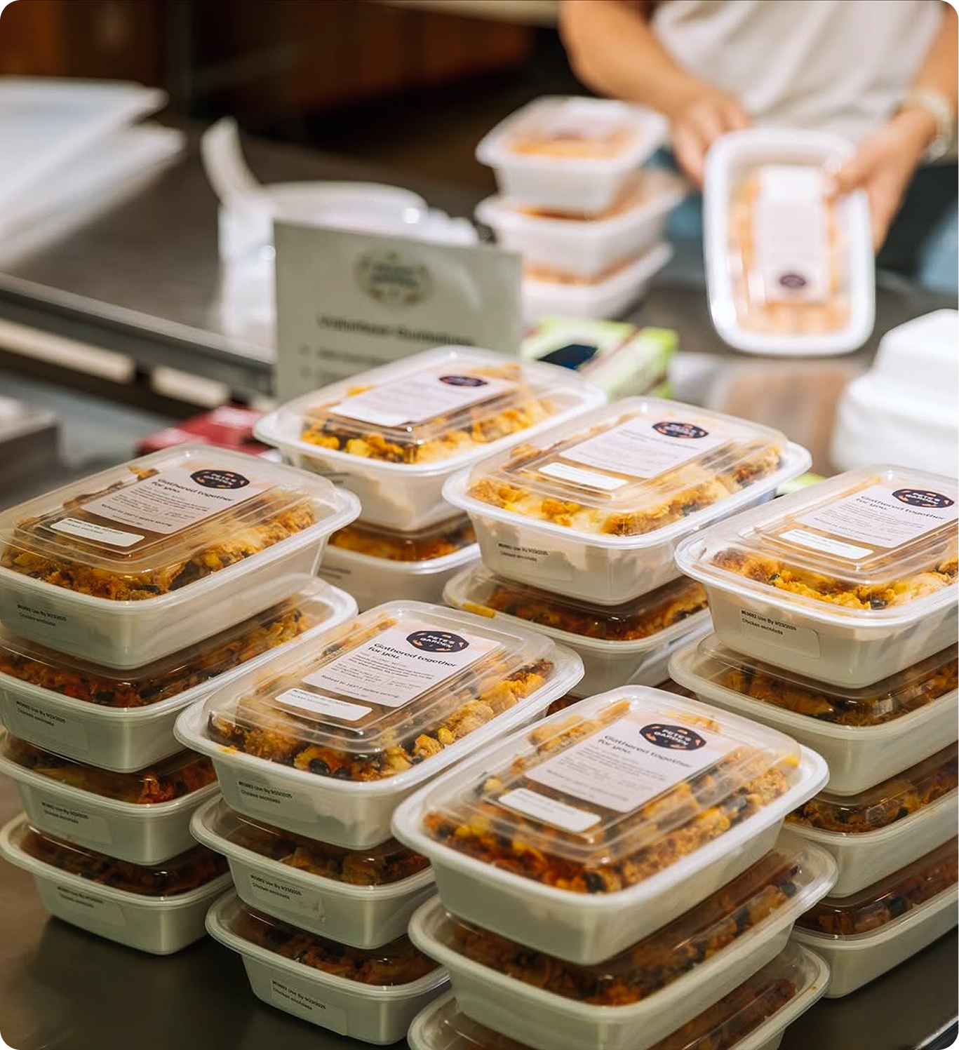 Stacks of pre-packaged meal containers on a commercial counter, with hands in the background preparing additional items.