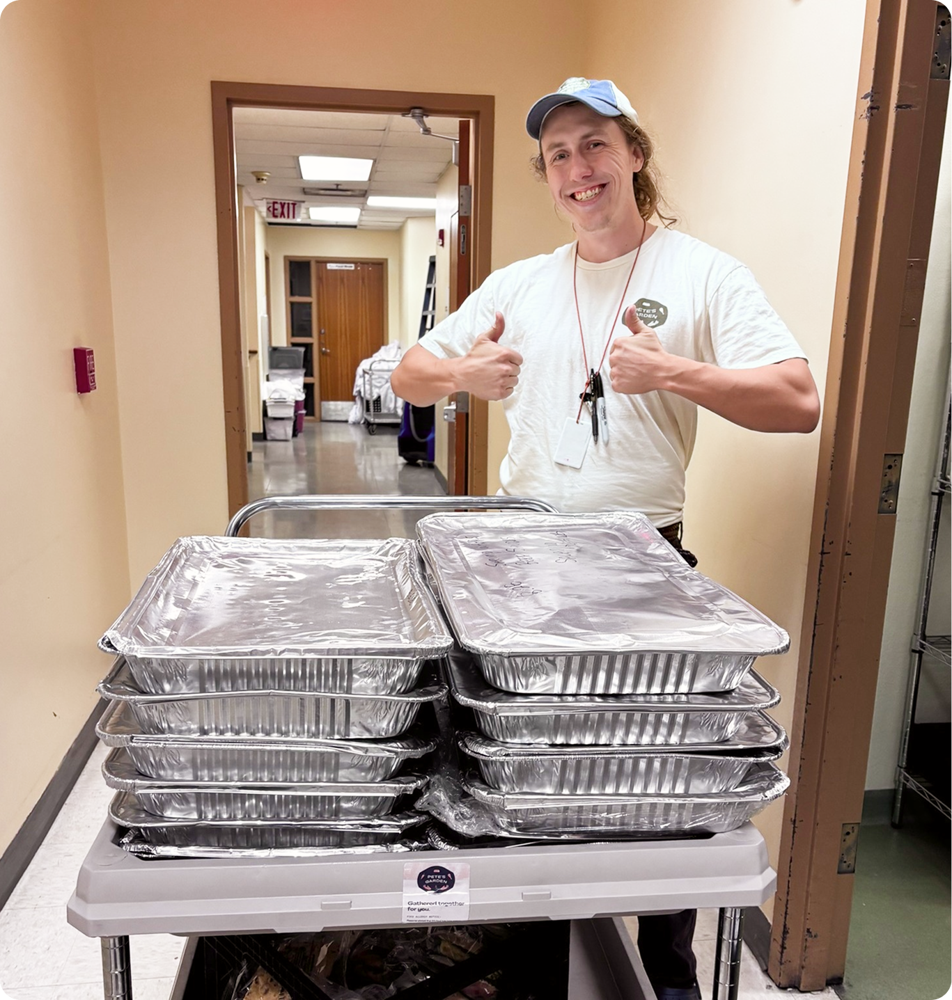 A person in a baseball cap smiles and gives two thumbs up while standing behind a cart stacked with aluminum food trays.