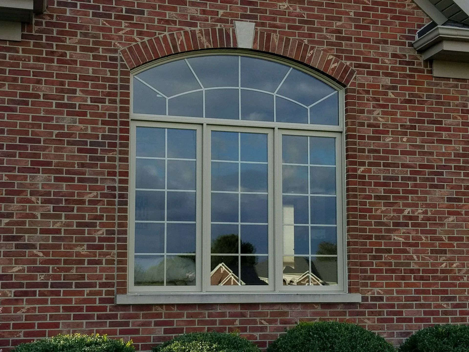A rectangular window with a curved top and grid muntins, set into a red brick wall above green shrubs.