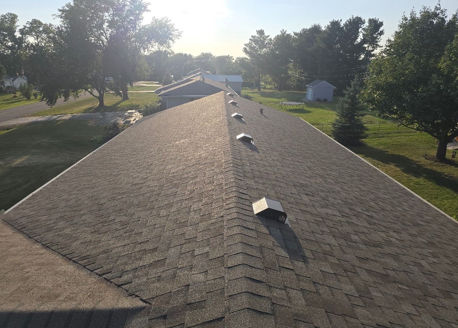 An aerial view of a gray asphalt shingle roof with several small ventilation caps, set against a sunny, tree-lined yard.
