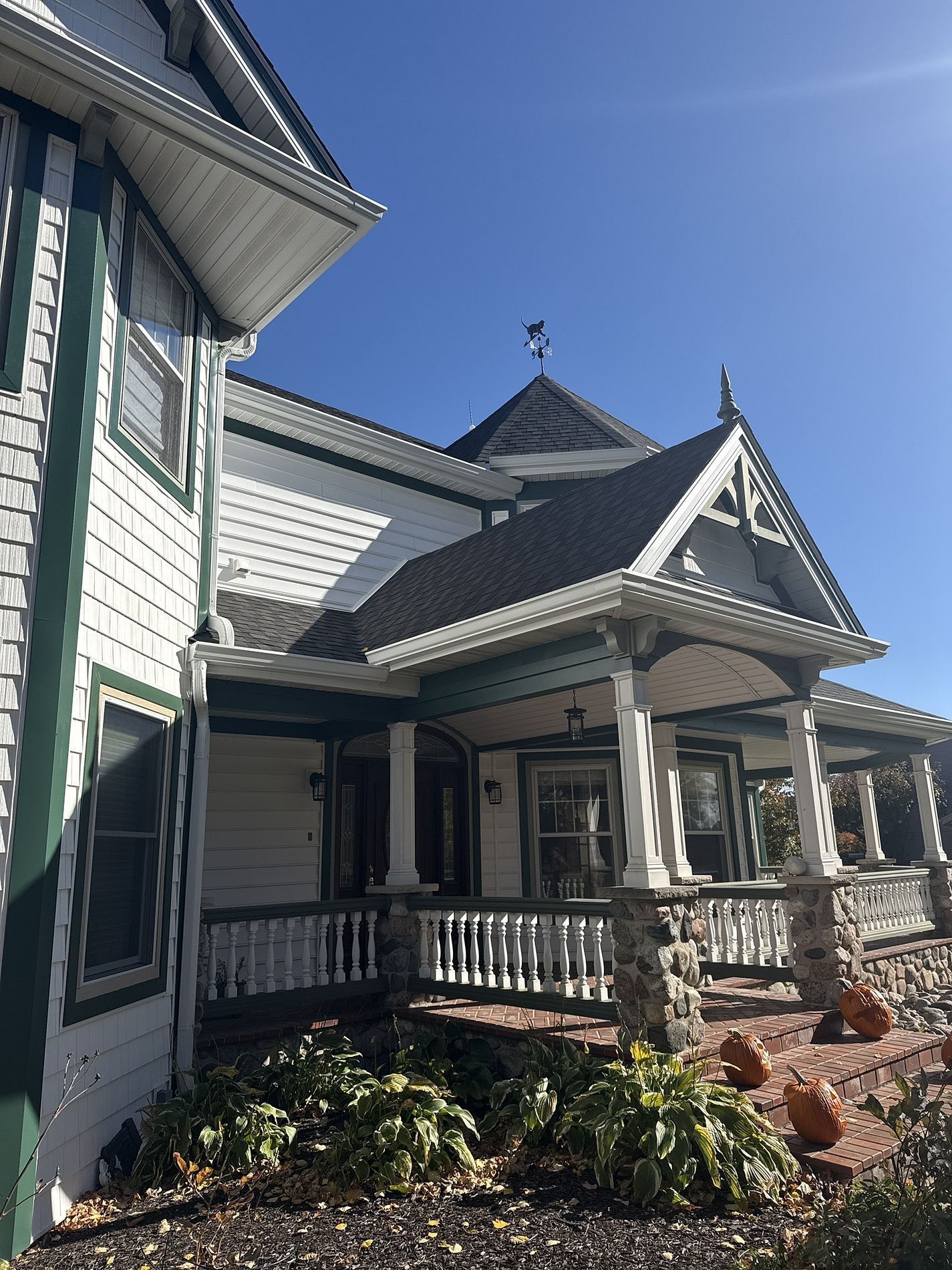 A two-story, white Victorian house with green trim, a large front porch with stone pillars, and a pointed roof cupola.
