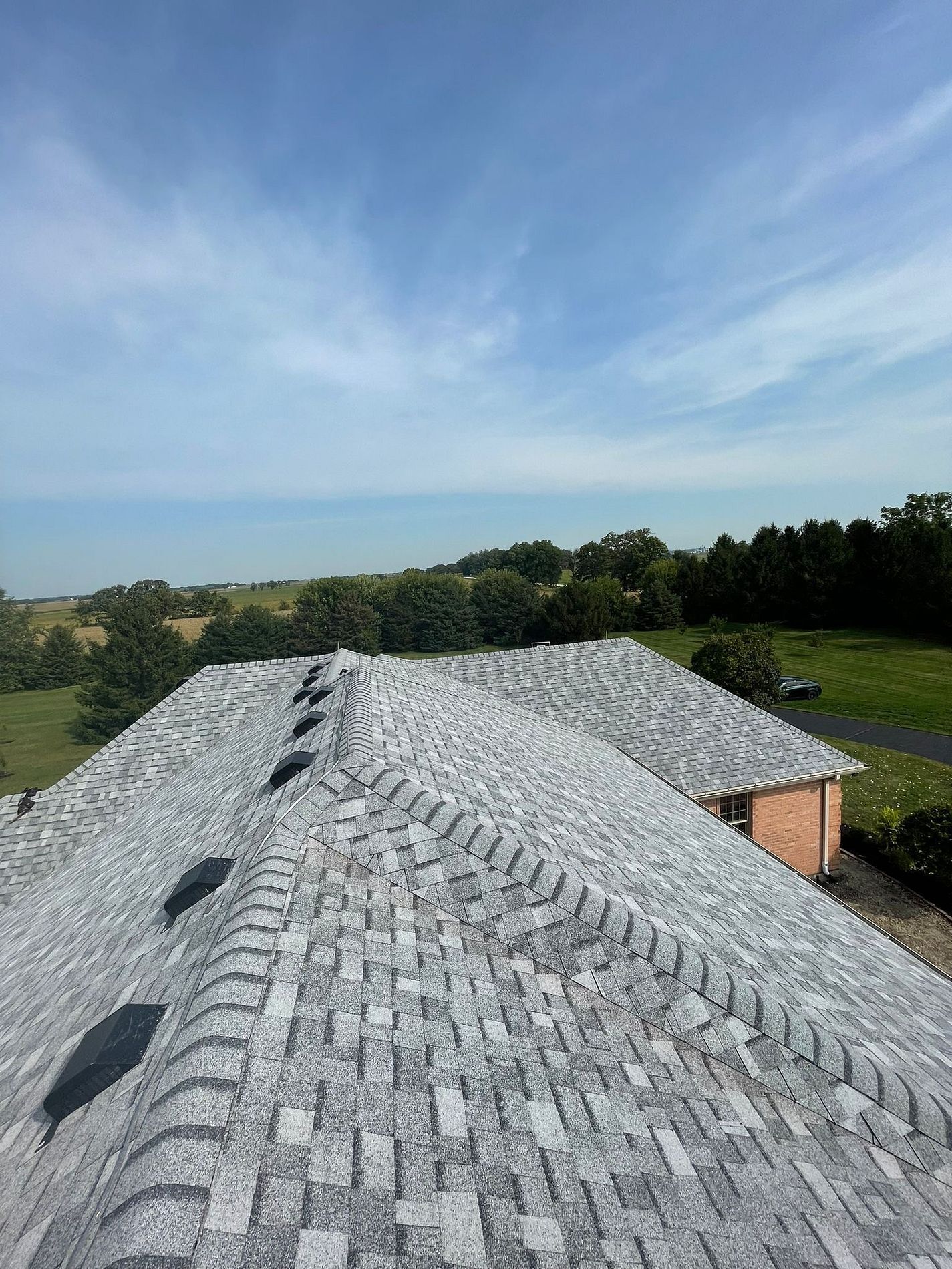 High-angle view of a gray shingled roof with several vents, overlooking a rural landscape with trees under a blue sky.