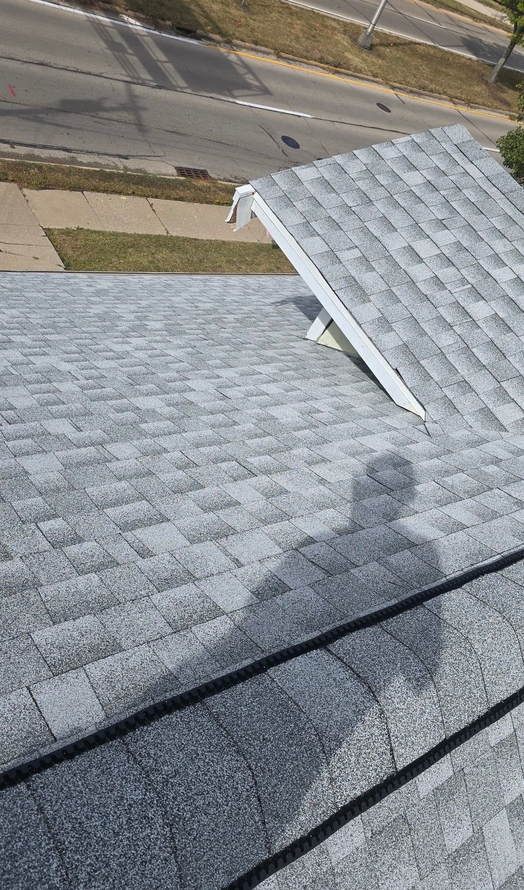 High-angle view of a grey shingled roof with a smaller dormer roof section, with a person's shadow cast upon it.