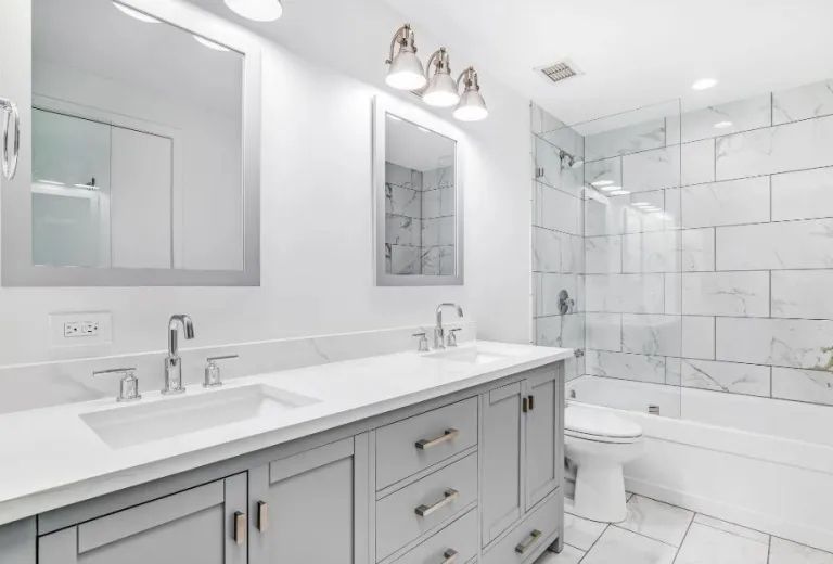 A modern bathroom with a light grey vanity, double sinks, white marble countertops, and a white tile bathtub enclosure.