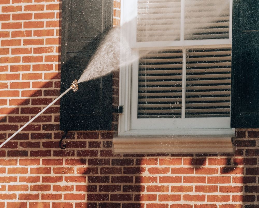 A pressure washer sprays water against a window and black shutter on a red brick house.