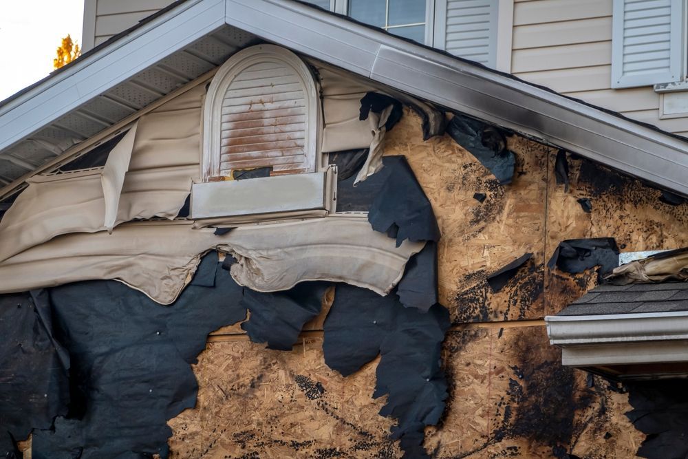 Damaged house exterior with torn siding, exposed wood sheathing, and shredded black weather barrier under a gable roof.