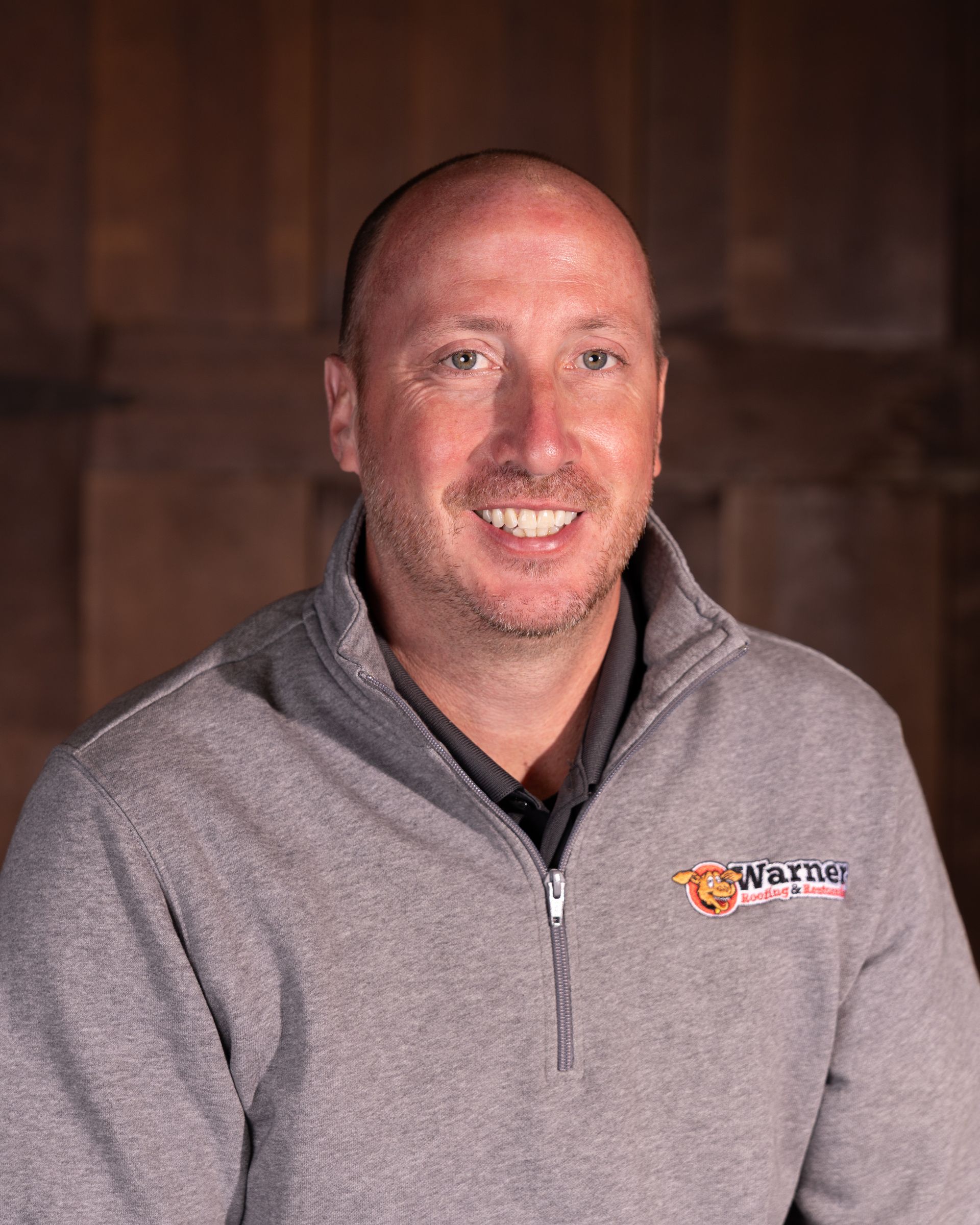 A person wearing a grey quarter-zip sweater with a logo, smiling in front of a wood-paneled wall.