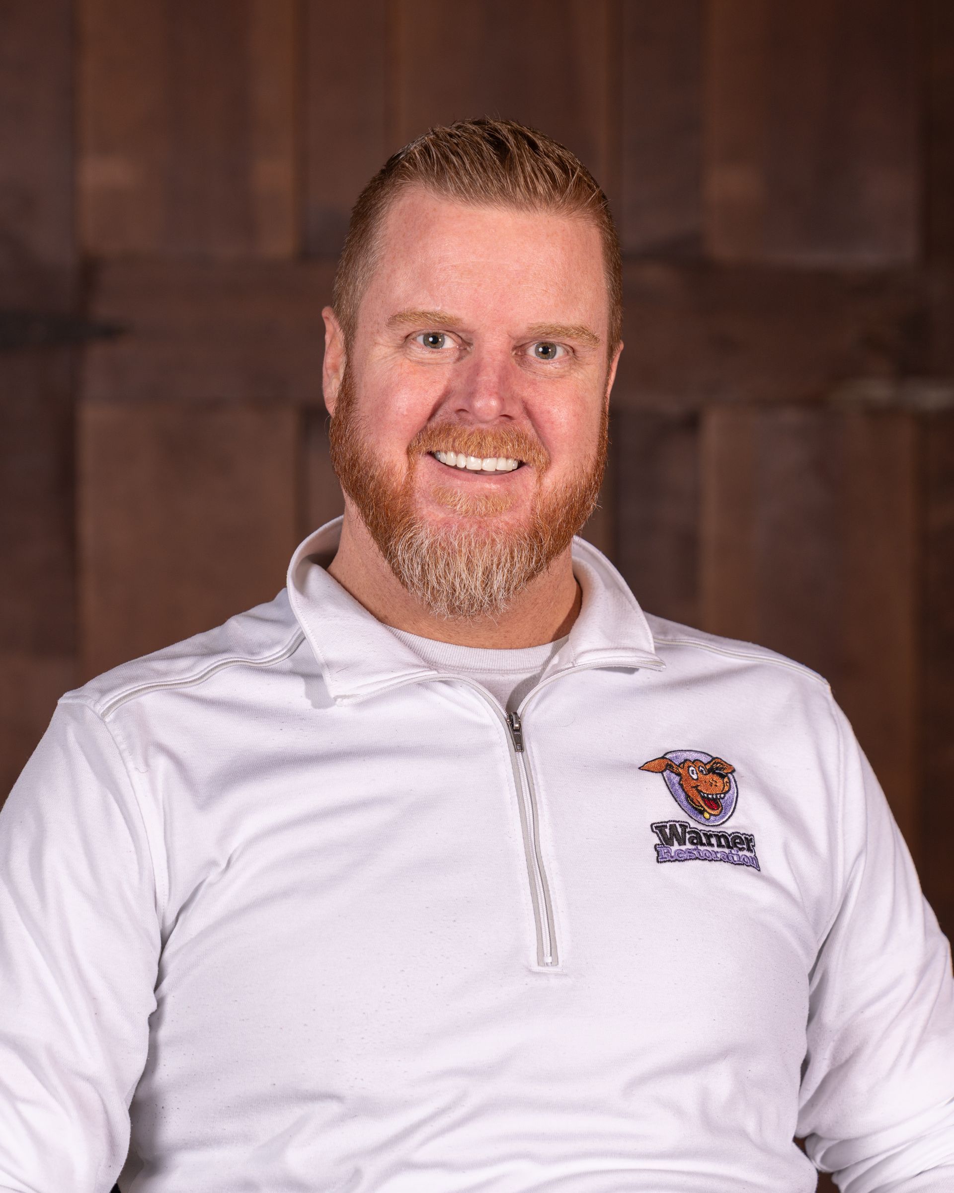 A smiling man with a reddish beard, wearing a white long-sleeve quarter-zip shirt with a logo, against a wood background.
