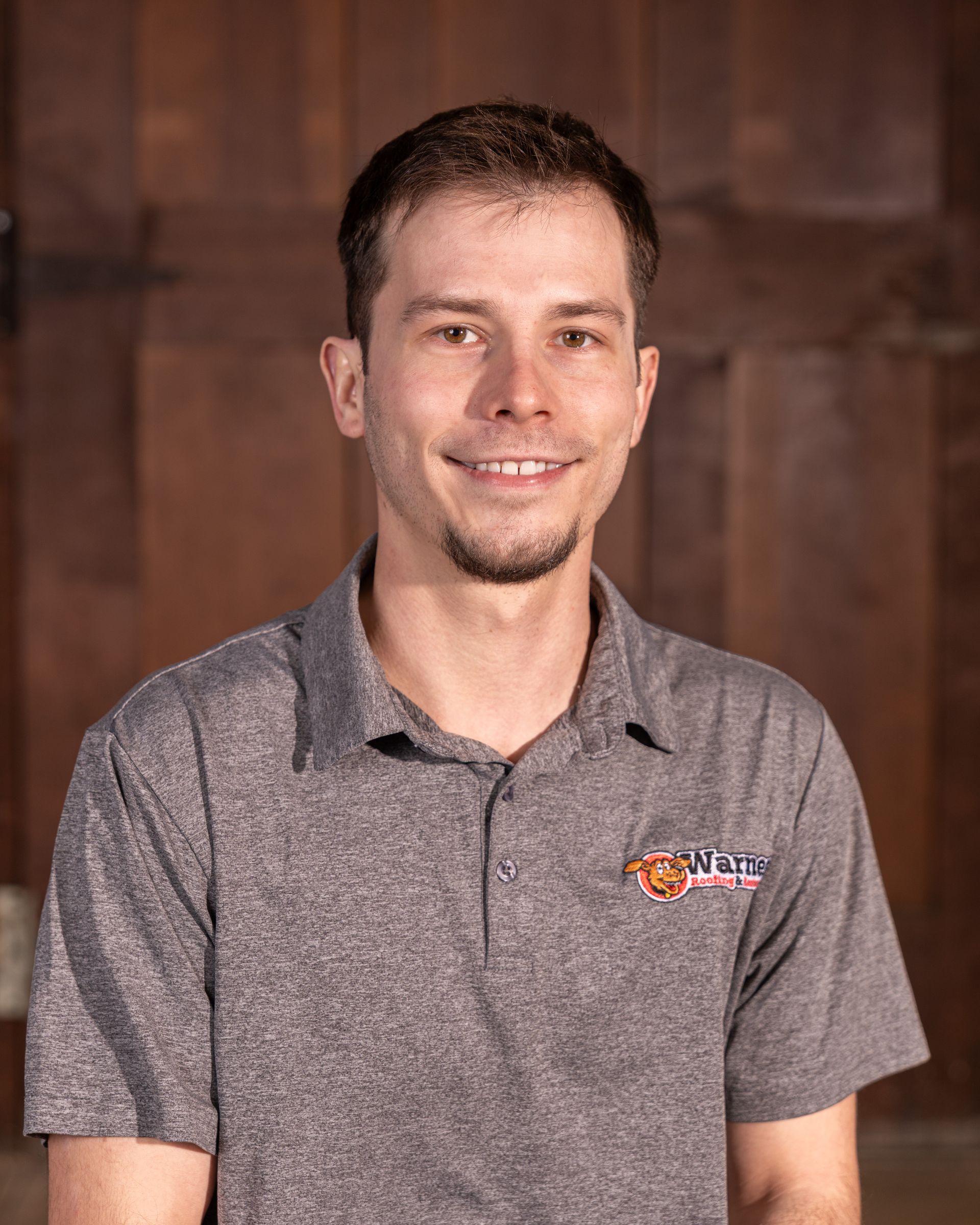 Smiling person wearing a gray polo shirt with a logo, positioned in front of a wood-paneled background.