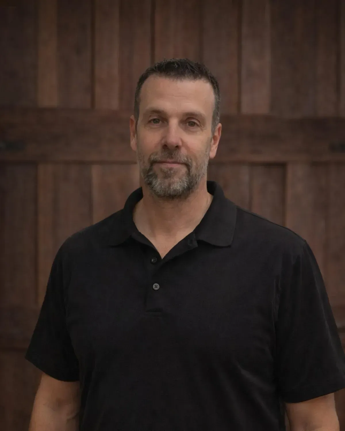 A man with a short beard wearing a black polo shirt, standing in front of a rustic wooden door.