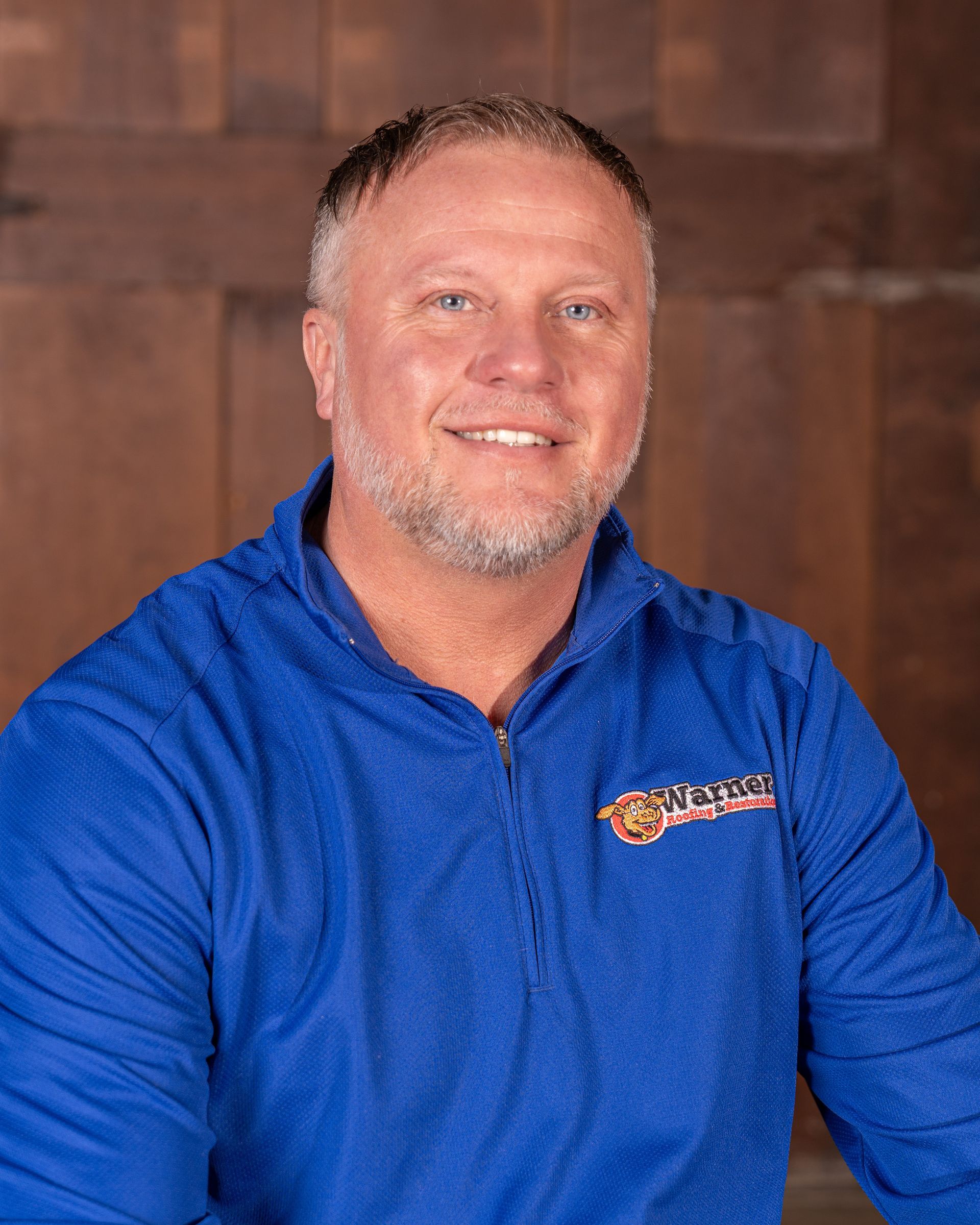 A smiling individual wears a blue long-sleeved quarter-zip shirt against a wood-paneled background.