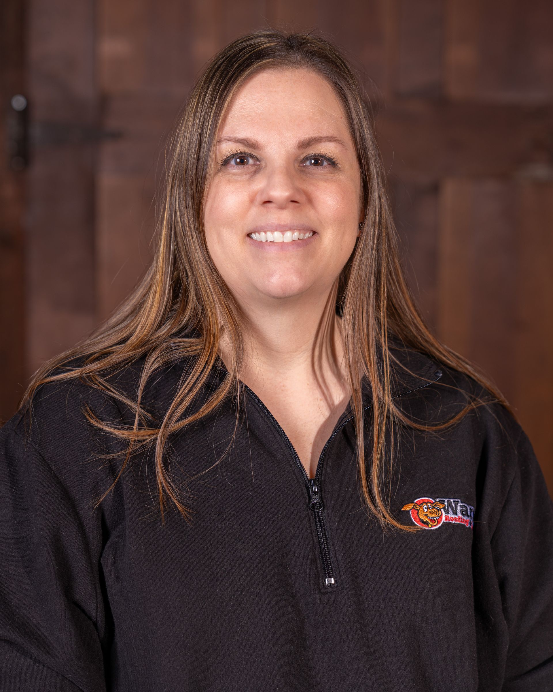 A person with long brown hair, wearing a black quarter-zip fleece, smiling against a wood-paneled background.