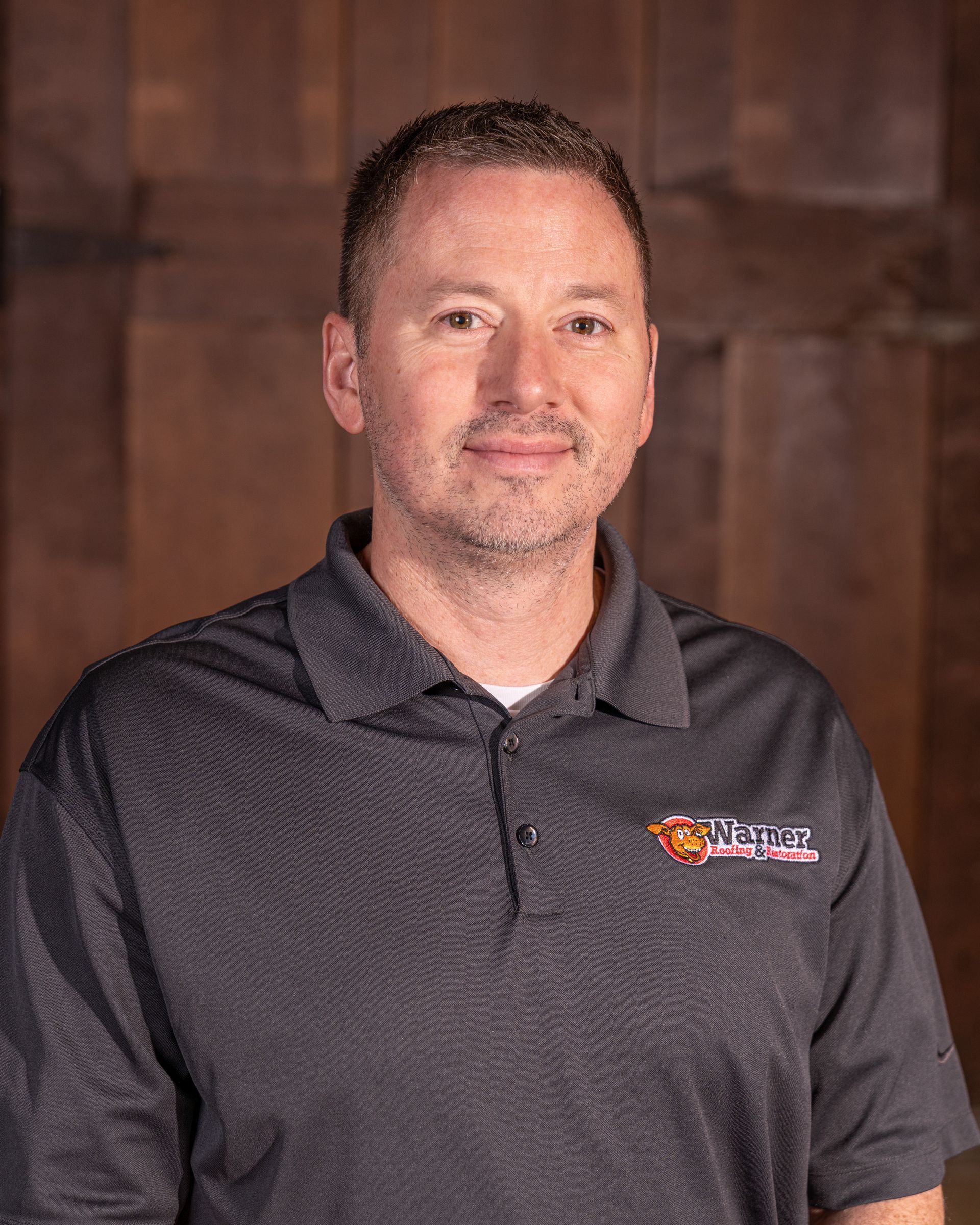 A person wearing a grey polo shirt with a logo, standing against a brown, vertical-plank wooden background.