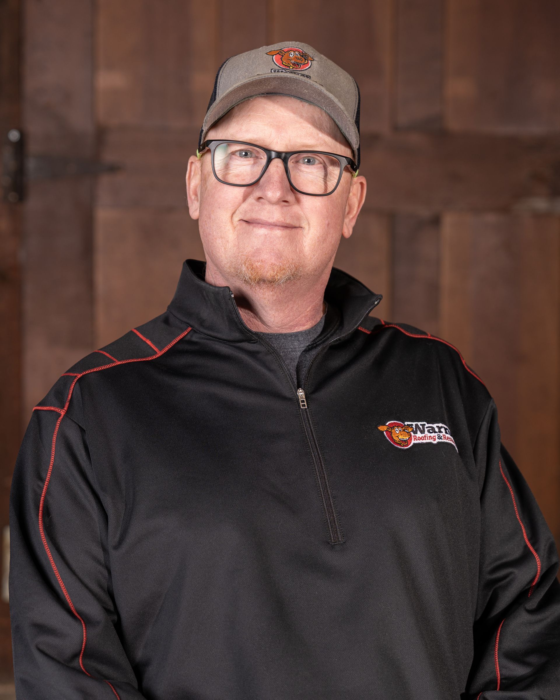A person wearing a grey cap and a black zip-up jacket with red accents, posing against a wooden door background.