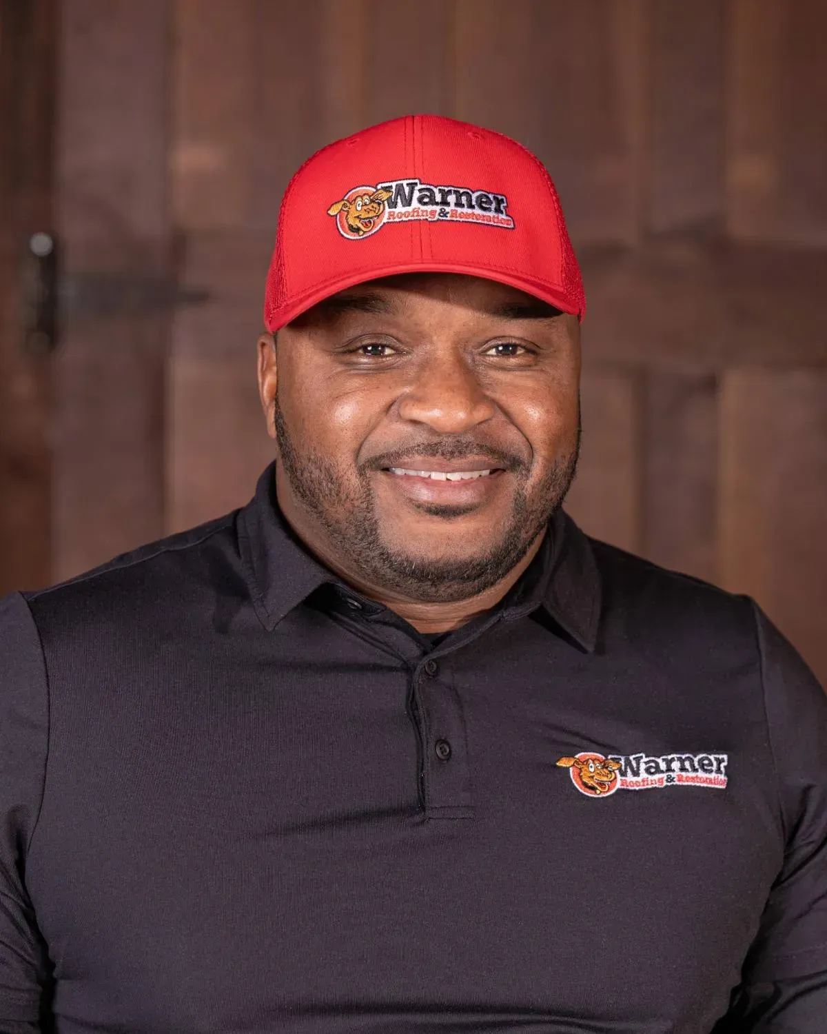 A smiling person wearing a red baseball cap and a black polo shirt, both featuring the Warner brand logo.