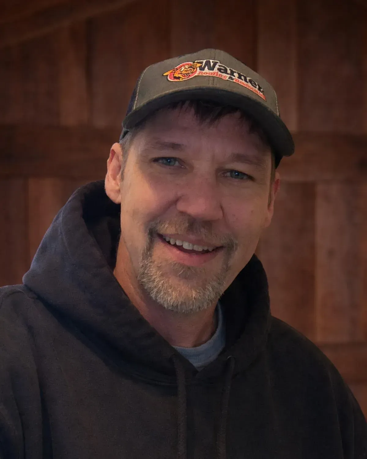 A man wearing a grey baseball cap and a black hooded sweatshirt smiles against a rustic, wooden-paneled background.