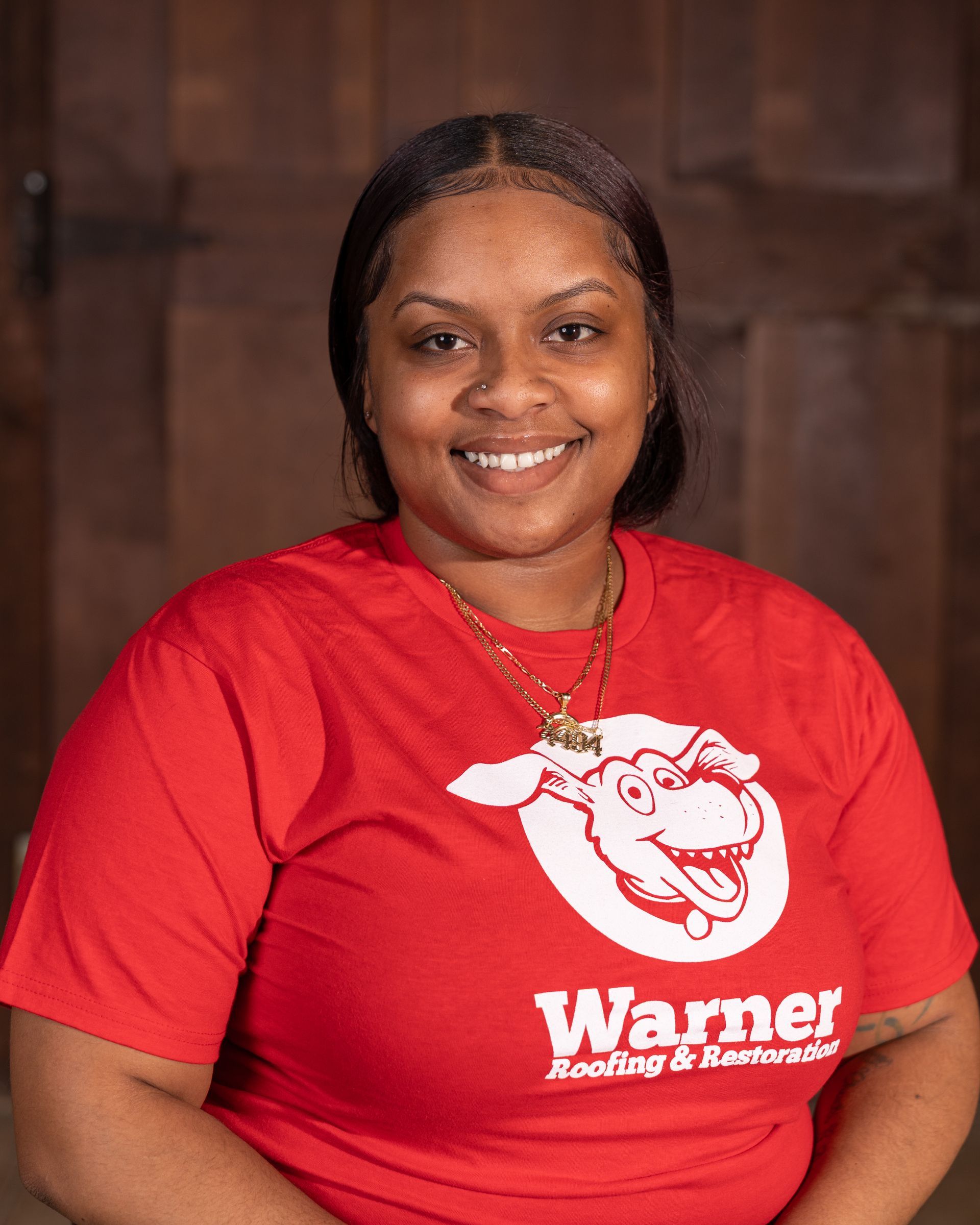 A smiling person wearing a red t-shirt with a logo and white text stands before a brown wood-paneled wall.