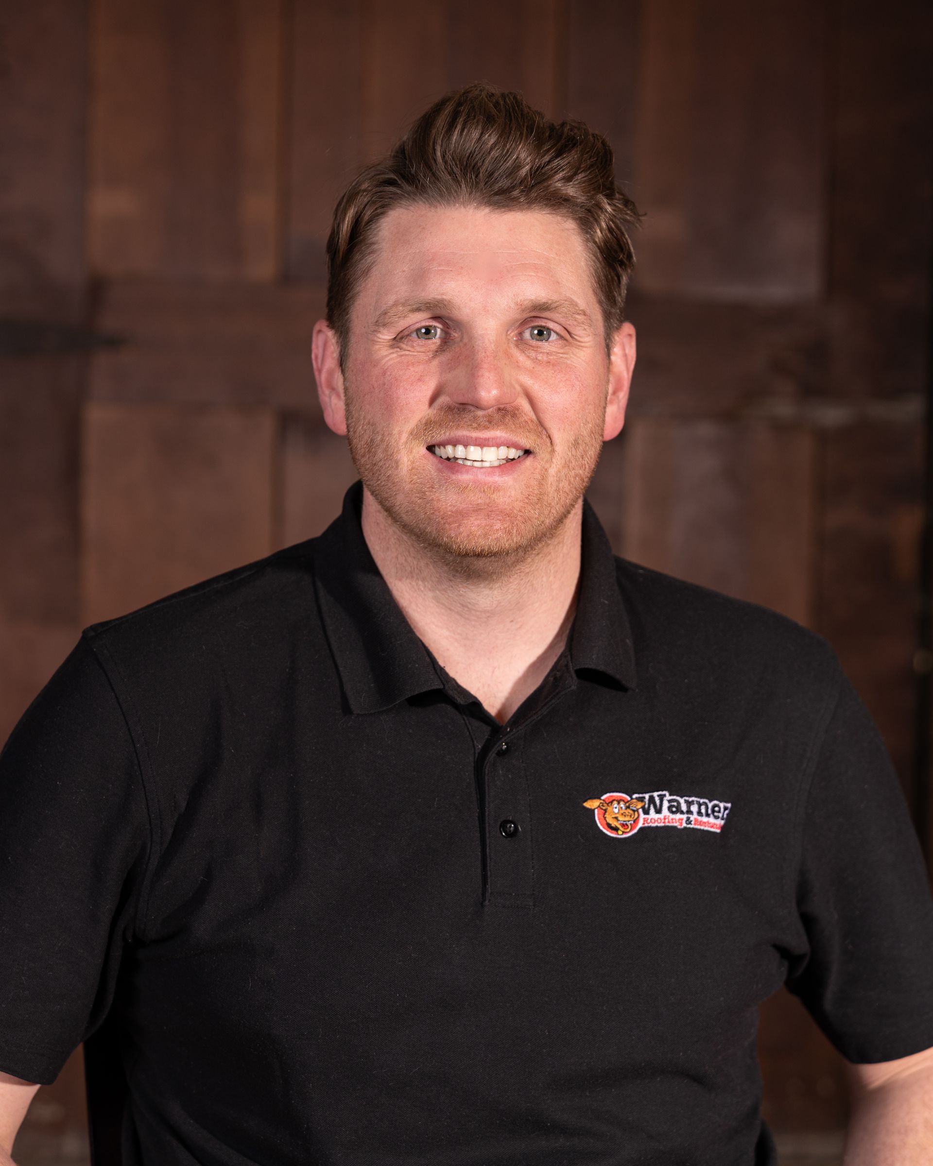 A smiling person wearing a black polo shirt with a logo, set against a brown wood-paneled background.
