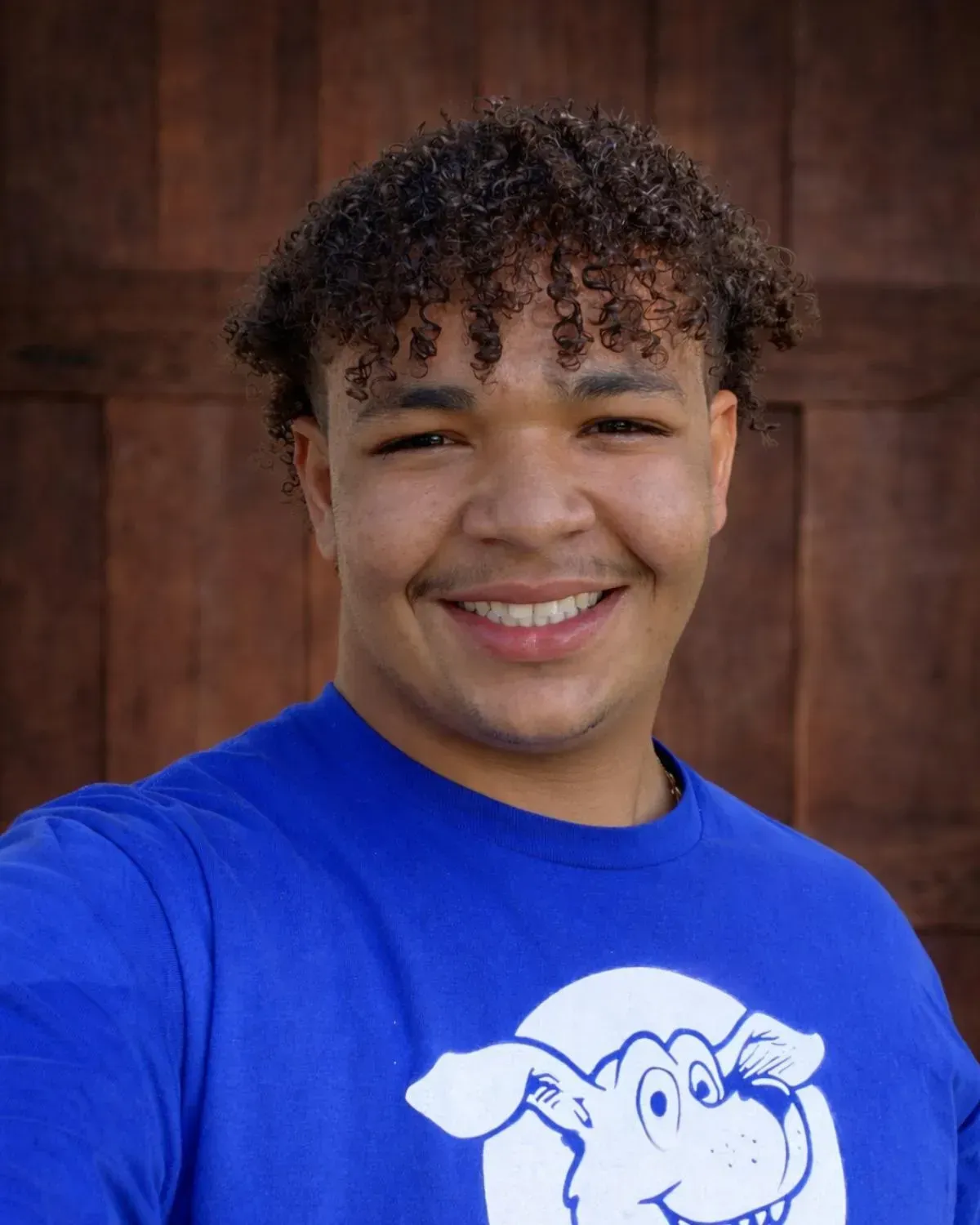 A smiling person with curly hair wears a blue t-shirt featuring a dog graphic, posed against a dark wood background.