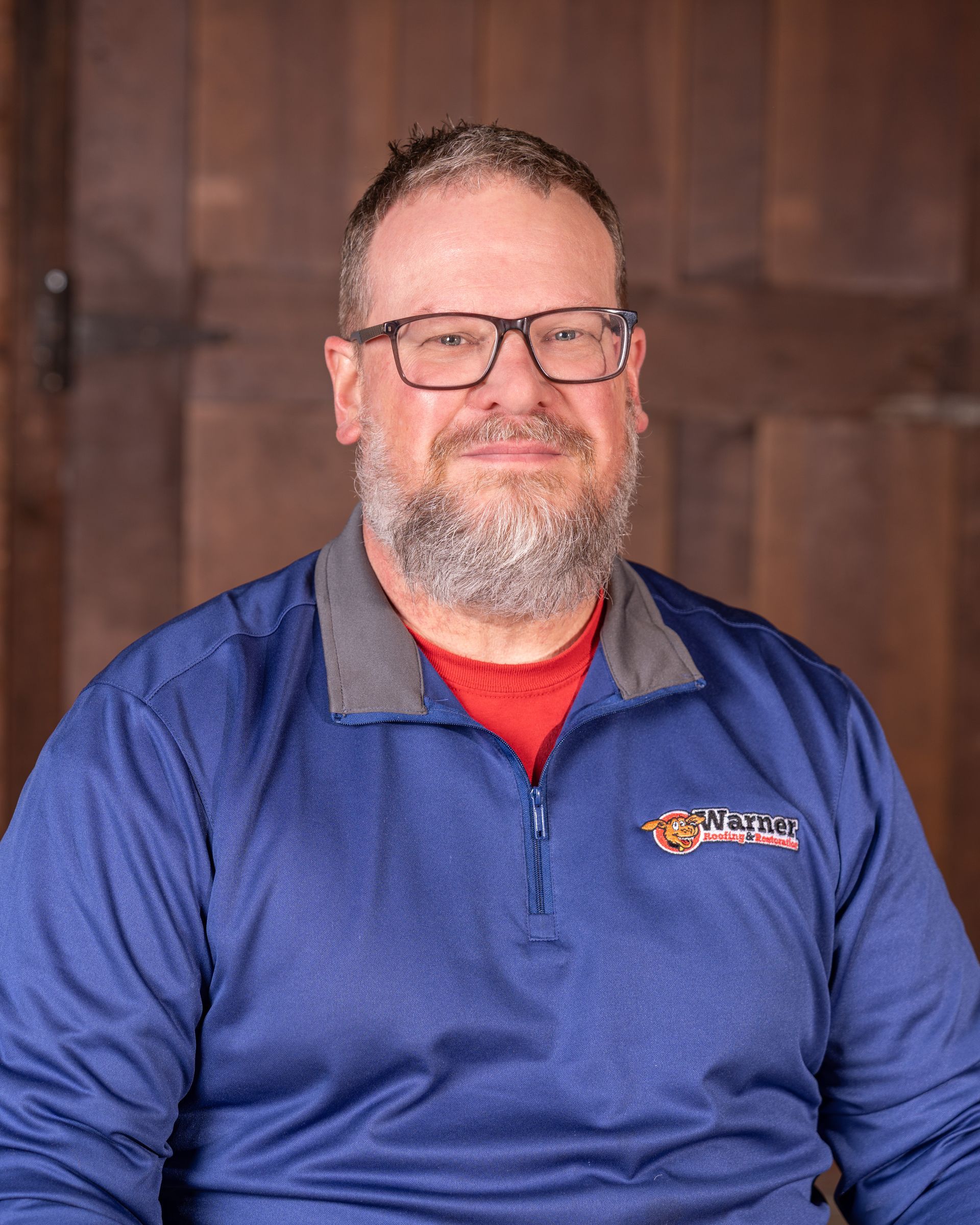 A person with a beard and glasses wearing a blue quarter-zip pullover over a red shirt against a wooden background.