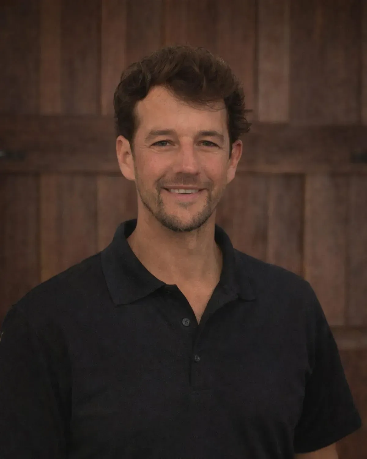 A person with short, dark curly hair and a light beard smiles, wearing a dark polo shirt against a wooden plank background.