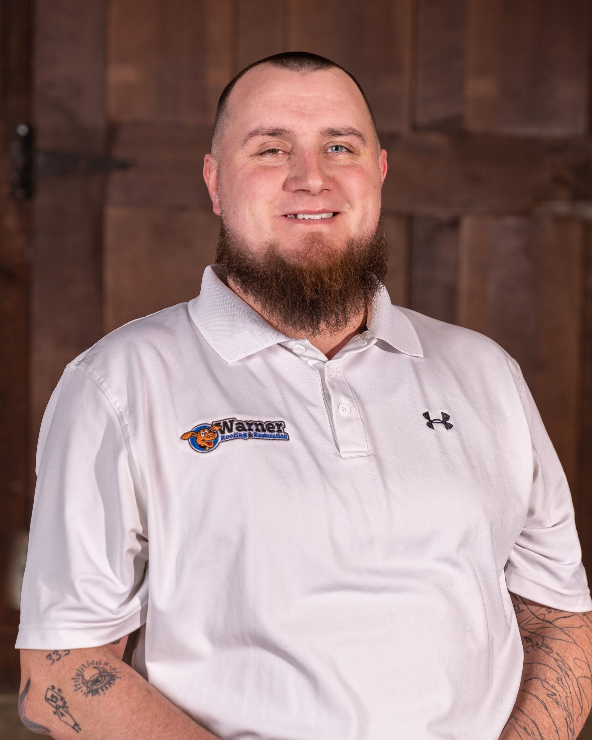 A person with a beard, wearing a white polo shirt with a logo, smiles against a rustic wood-plank background.