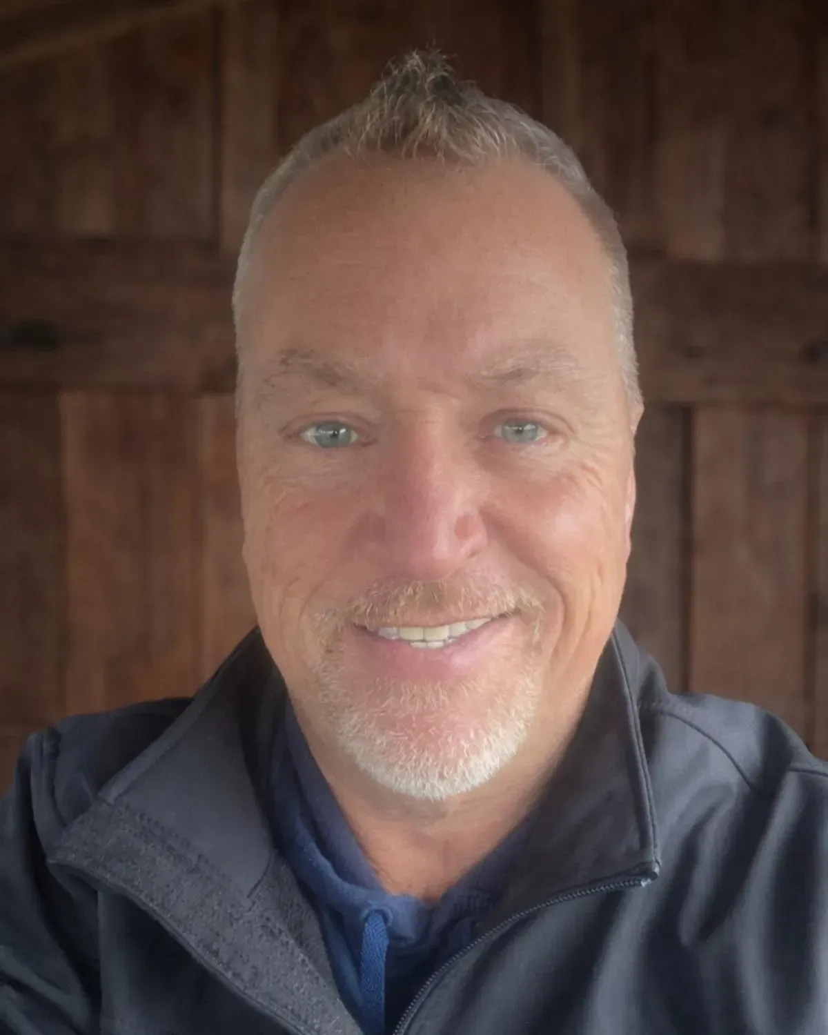 A person with short, light-colored hair and a goatee smiles in a selfie, wearing a dark jacket against a wood wall.